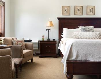 Two Tan chairs to the left and to the right is the Wooden California King bed with a white bedspread, night stand inbetween